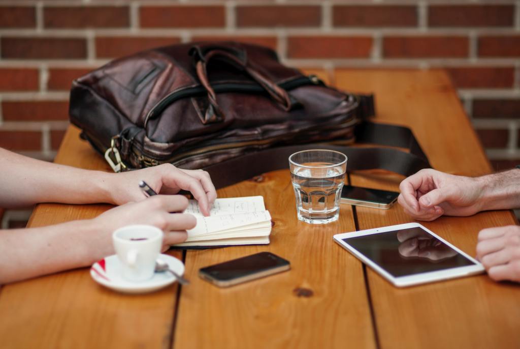 Photo shows hands, notebooks, and communication devices on a cafe table, indicating a work meeting over coffee. 